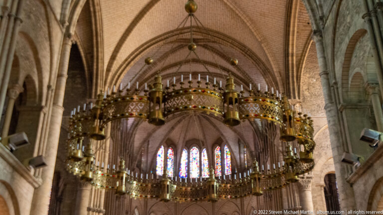 Large gilded circular corona chandelier suspended beneath church vaults, with many candle holders and miniature towers.