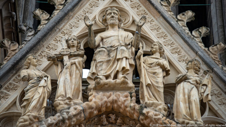 Seated Christ figure raising one hand in blessing, flanked by standing figures in a pointed Gothic gable at Reims Cathedral.