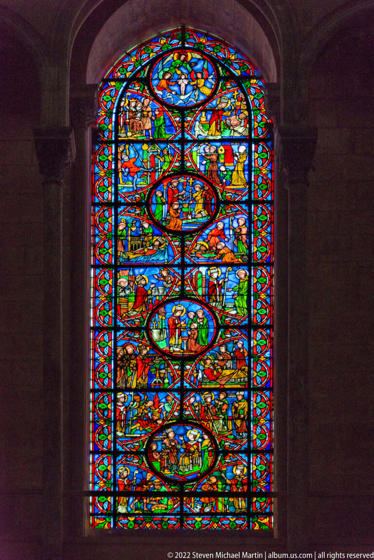 Single tall stained-glass lancet filled with stacked narrative medallions and multicolored ornamental borders.