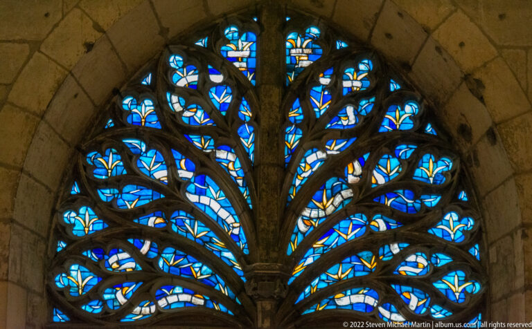 Pointed window with branching tree-like stone tracery and blue stained glass patterned with small motifs.