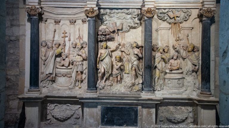 Three stone relief panels showing clerics, crosses, altars, and ceremonial scenes between dark columns.