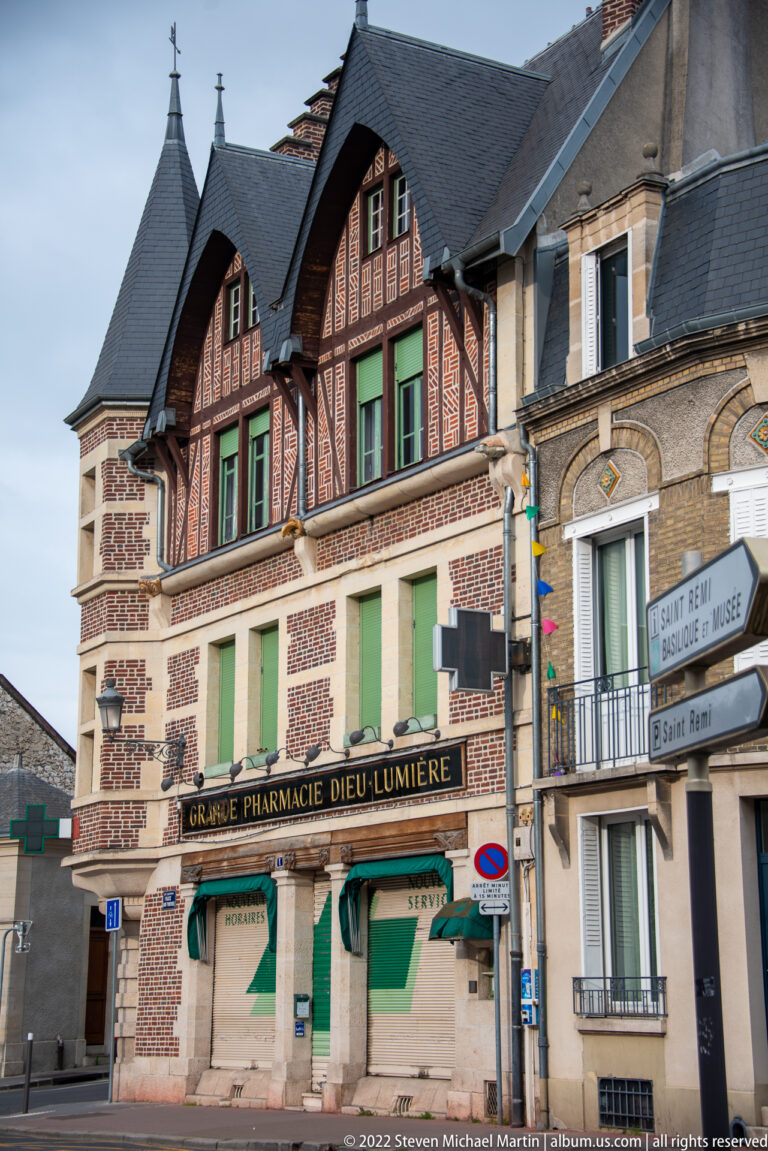 Decorative brick and half-timbered corner building marked “Grande Pharmacie Dieu-Lumière,” with green shutters and slate roofs.