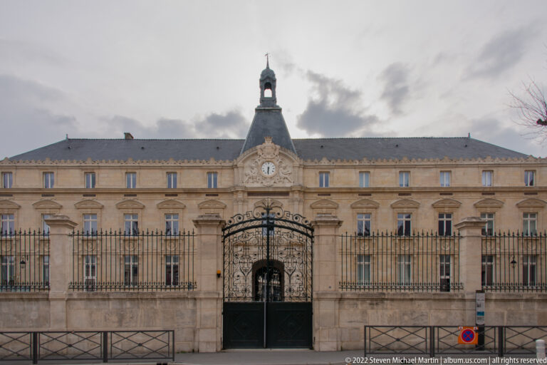 Symmetrical classical building with ornate iron gates, central clock, small cupola, and rows of tall windows.