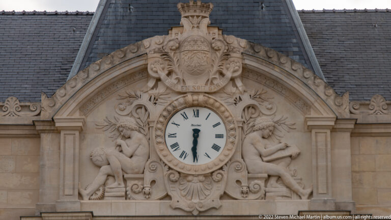 Round Roman-numeral clock framed by carved scrollwork, two reclining figures, and a heraldic crest.