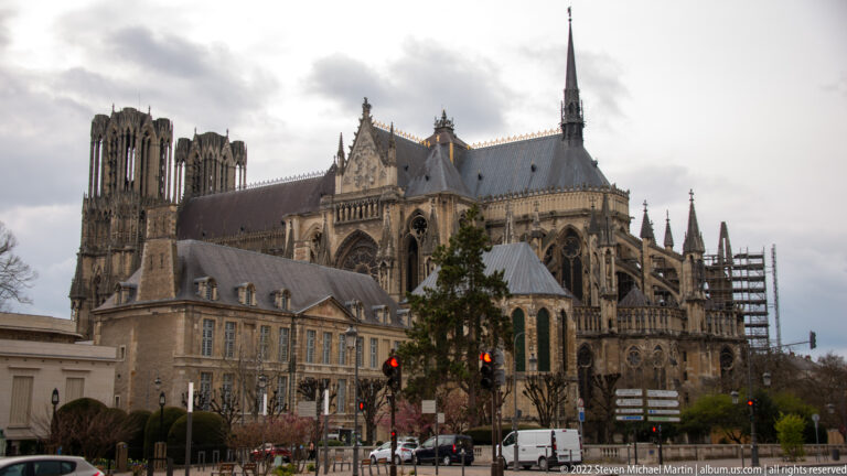 Side and apse view of Reims Cathedral with flying buttresses, central spire, distant west towers, and scaffolding.