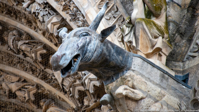 Dark animal-headed gargoyle with open mouth and metal support projecting from richly carved stonework at Reims Cathedral.
