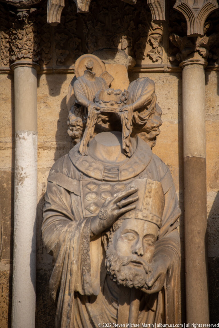 Headless stone saint or bishop holding a mitred severed head between architectural columns.
