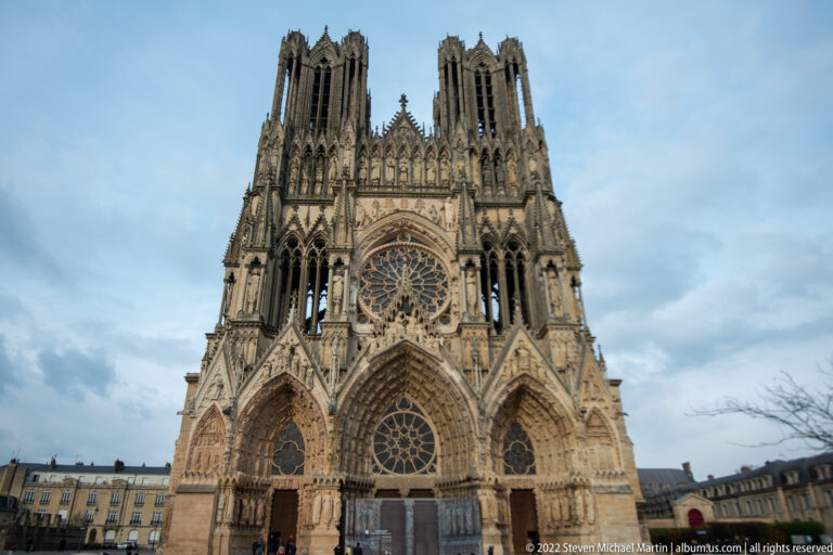 Full west façade of Reims Cathedral with three portals, central rose window, dense sculpture, and twin towers.