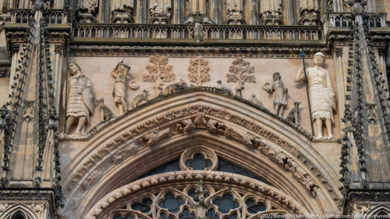 Standing and smaller carved figures with trees arranged in a horizontal register above a pointed cathedral portal.