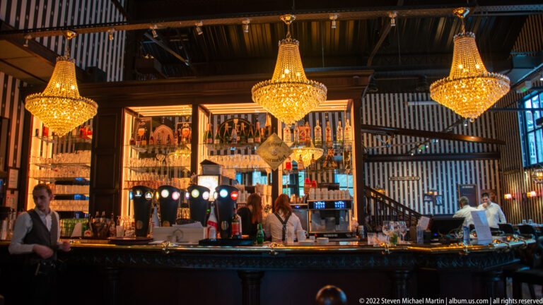 Contemporary bar with three crystal chandeliers, mirrored bottle shelves, beer taps, and staff behind the counter.