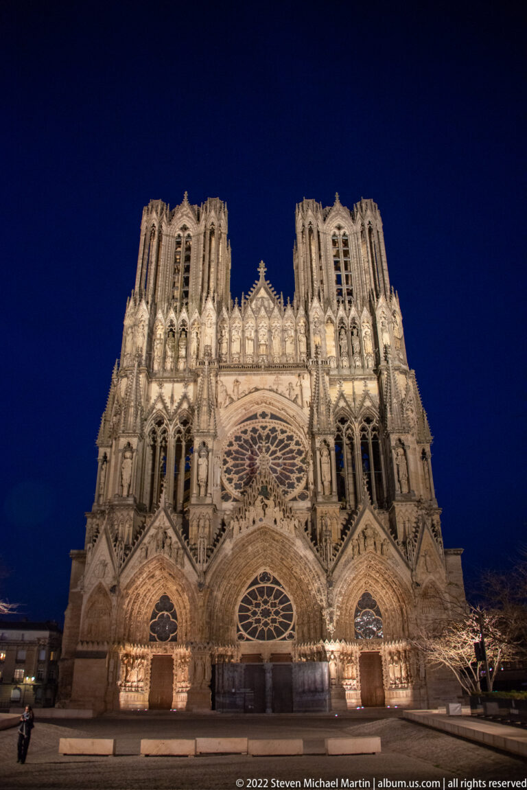 Illuminated Reims Cathedral west façade at night beneath a deep blue sky.