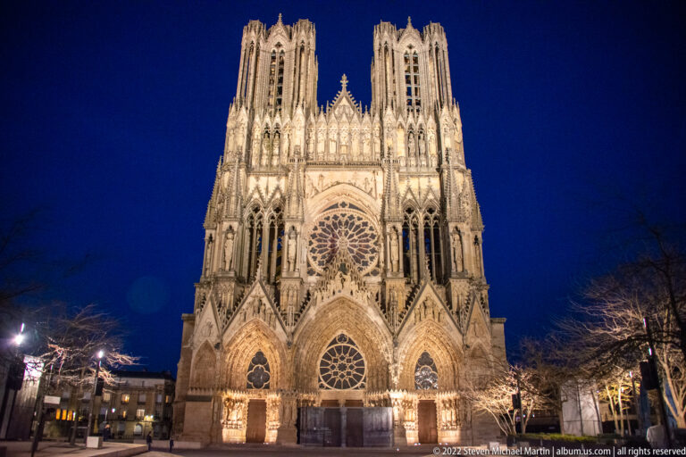 Frontal night view of Reims Cathedral with illuminated twin towers, rose window, and three west portals.