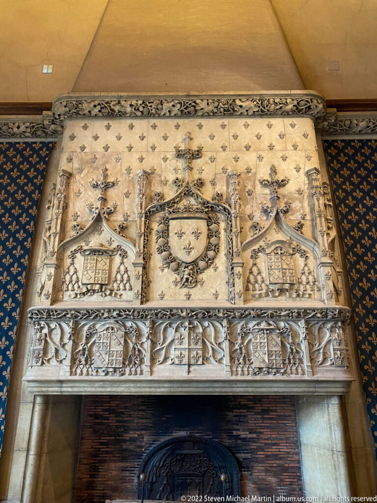 Ornate stone fireplace with crowned fleur-de-lis shield, heraldic reliefs, and blue fleur-de-lis wall covering.