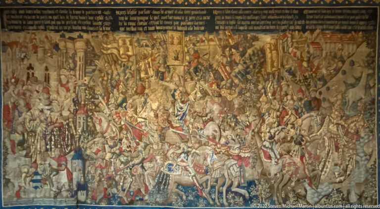 Large woven battle scene crowded with cavalry, infantry, banners, and distant architecture beneath a text band.