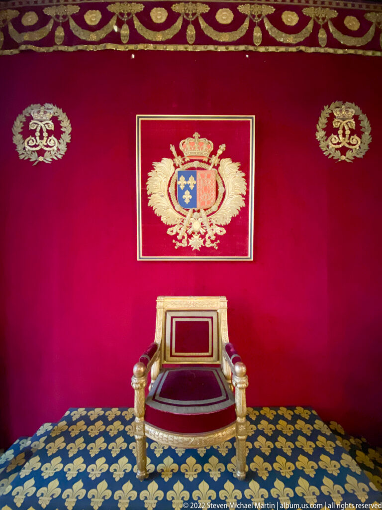 Red and gilt ceremonial throne beneath a royal coat of arms on a red wall over fleur-de-lis carpet.