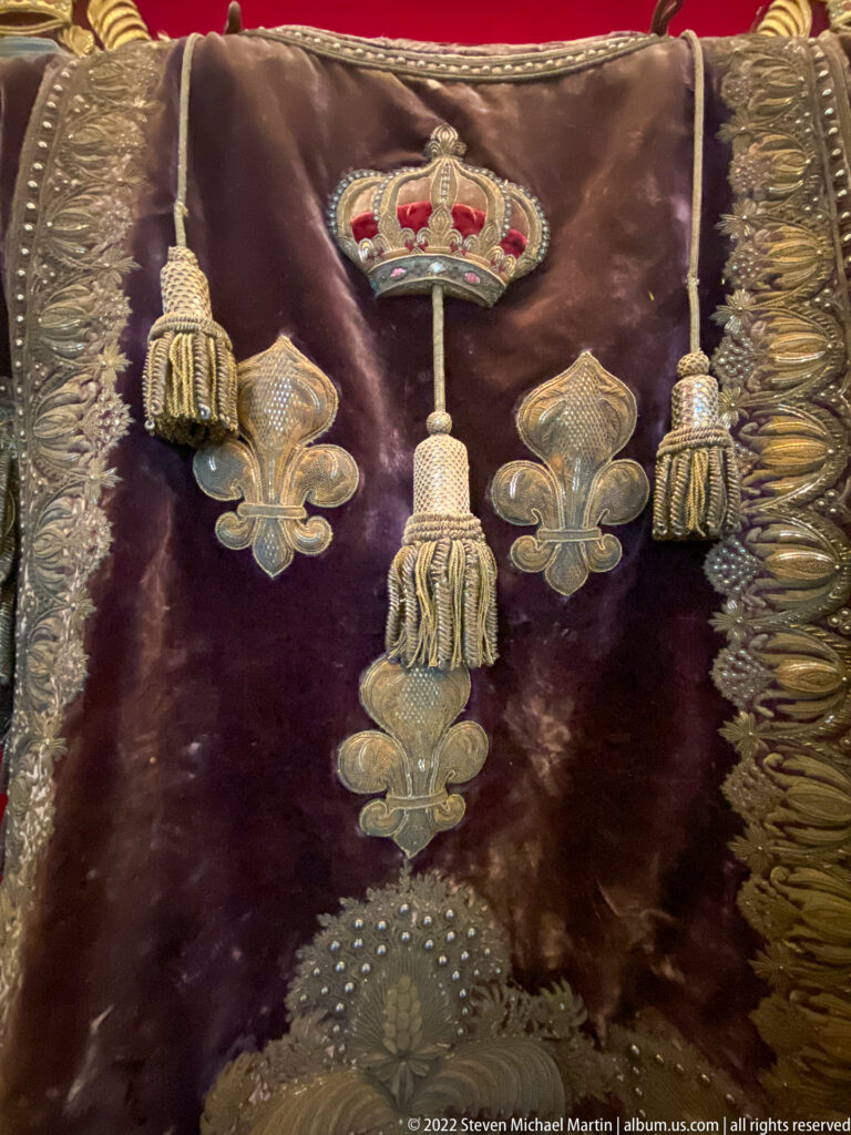 Dark velvet textile with small crown, fleur-de-lis motifs, metallic tassels, fringe, and visible wear.