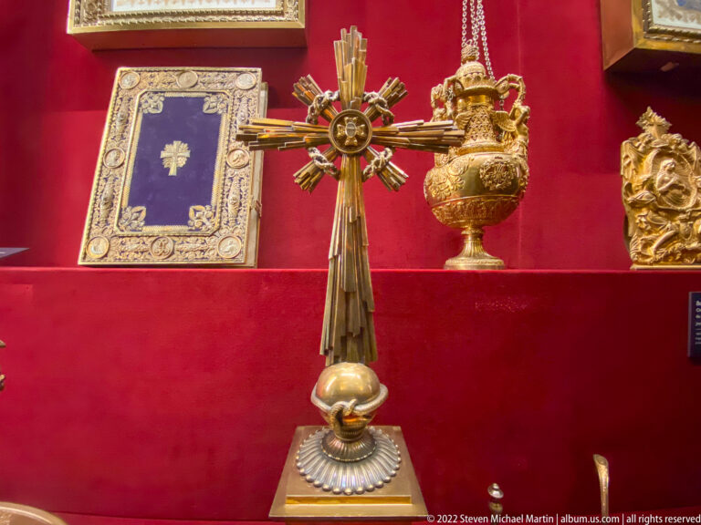 Processional cross on a pedestal beside a gilded vessel and ornate framed object against red museum fabric.