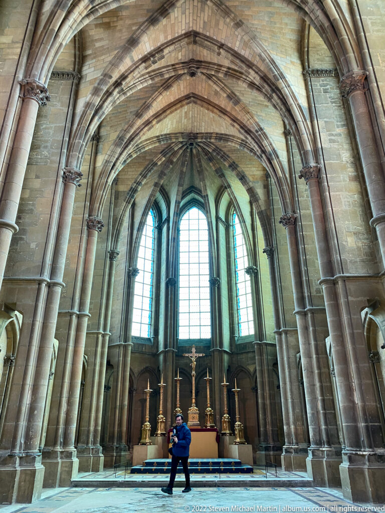 Small Gothic apse or chapel with rib vaults, tall lancet windows, altar, and one visitor.