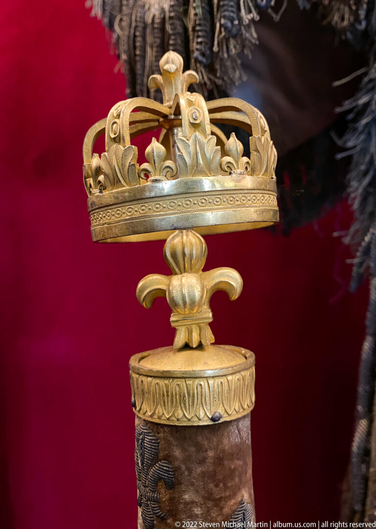 Gold crown-shaped finial with fleur-de-lis ornament mounted on a dark velvet-covered ceremonial shaft.