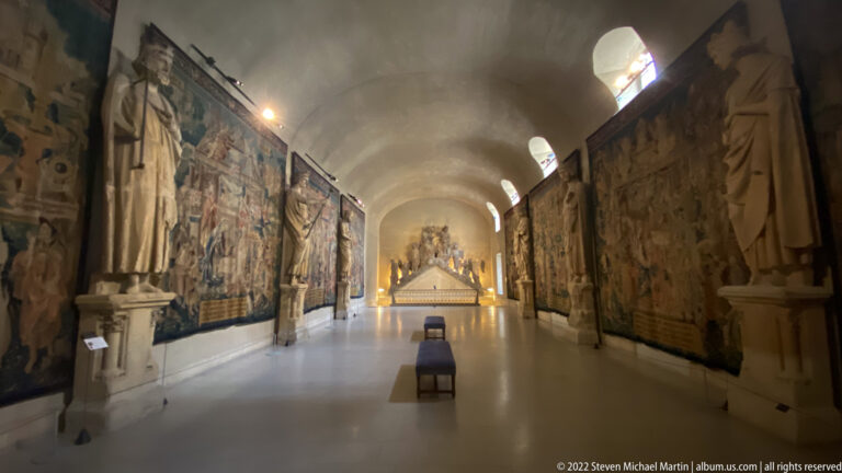 Long vaulted museum gallery with monumental tapestries, stone sculptures, high windows, and one chair on the centerline.