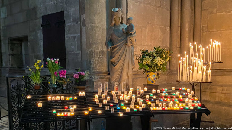 Virgin and Child statue above many votive candles and flowers, with tall burning tapers at right.