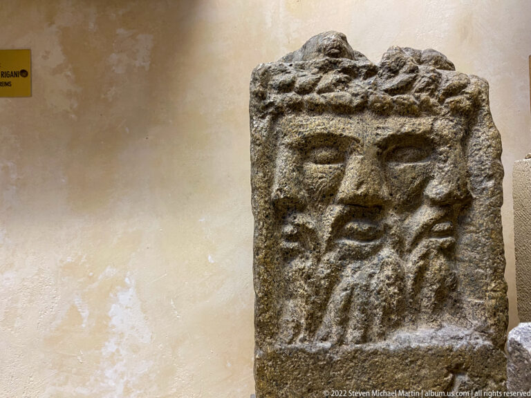 Rough stone block carved with a frontal bearded face or mask, with broken edges around the relief.