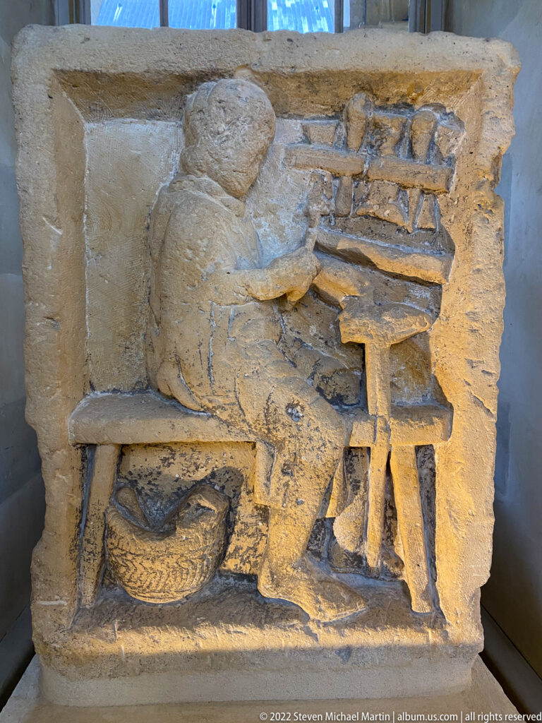 Stone relief of a seated worker operating a framed bench or table, with a basket beneath.
