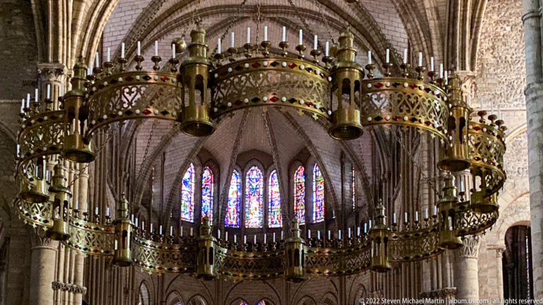 Large gilded circular corona chandelier with miniature towers, colored stones, and candles before stained-glass windows.