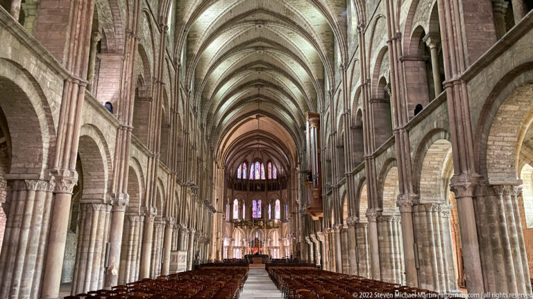 Long Saint-Remi nave with repeated stone bays, rib vaults, upper arcades, wooden chairs, and distant stained glass.