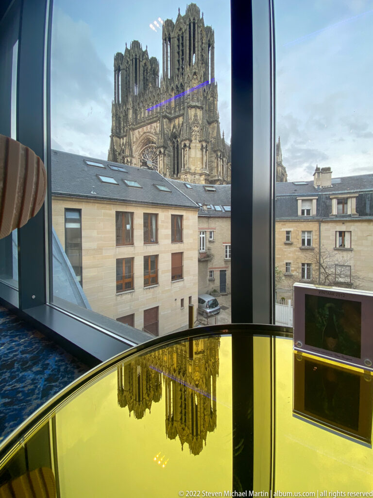 Reims Cathedral seen through a modern window above nearby roofs, reflected in a glossy yellow table.
