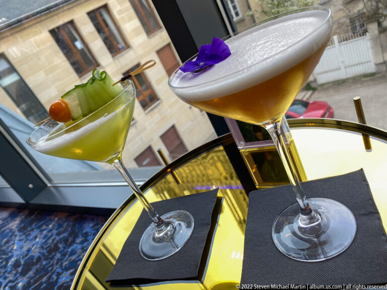 Green garnished cocktail and amber foamy cocktail in stemmed glasses on a glossy yellow table.