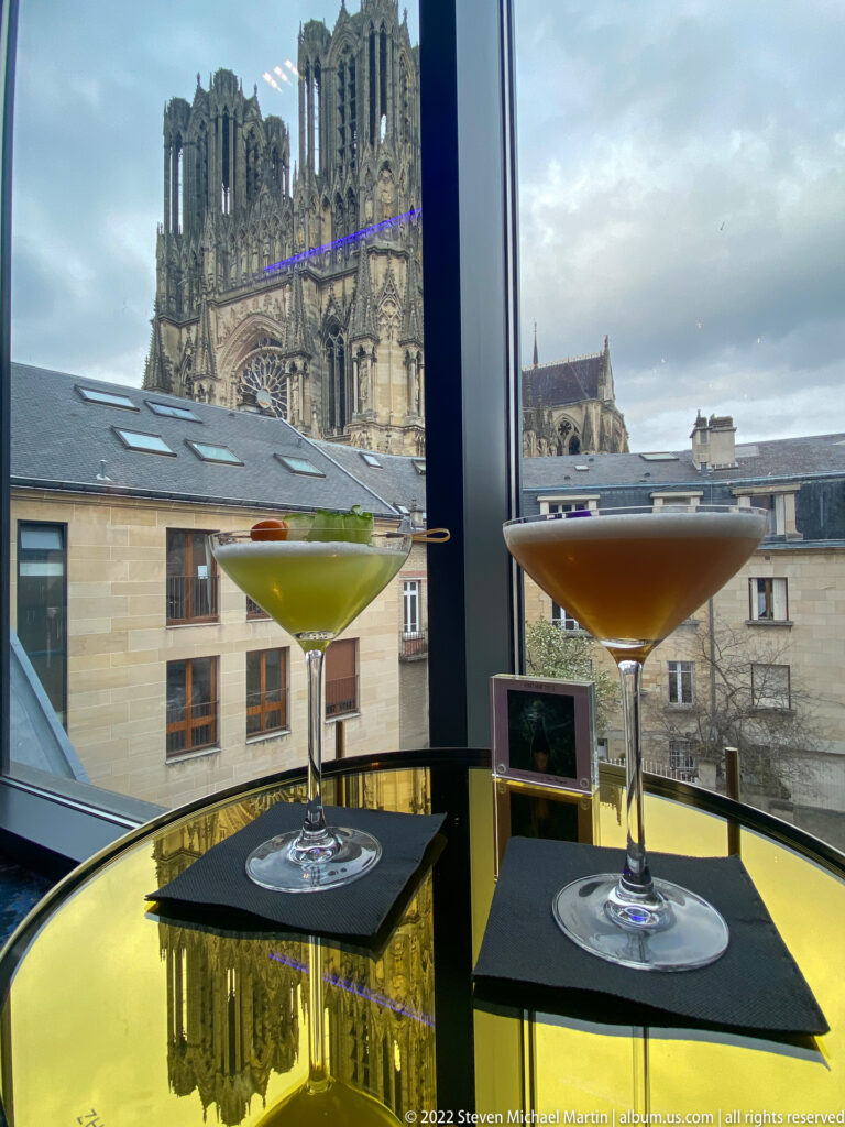 Two cocktails on a yellow table before a window view of Reims Cathedral and nearby rooftops.