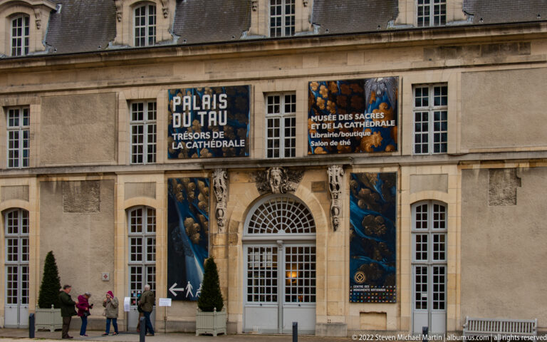 Palais du Tau entrance in Reims with blue museum banners, central arched doorway, and visitors on the pavement.