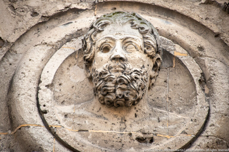 Weathered bearded male stone head projecting from a circular medallion on the Porte de Mars.