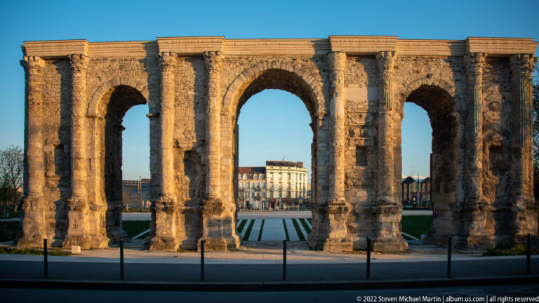 Frontal view of the three-arched Roman Porte de Mars in warm light, with city buildings visible through the openings.
