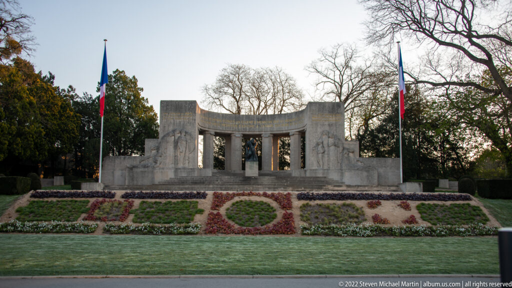Curved stone war memorial with central columns, French flags, and patterned flower beds in a formal garden.
