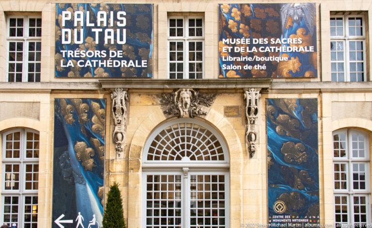 Close view of the Palais du Tau entrance with arched doorway, carved stone ornament, and blue museum banners.