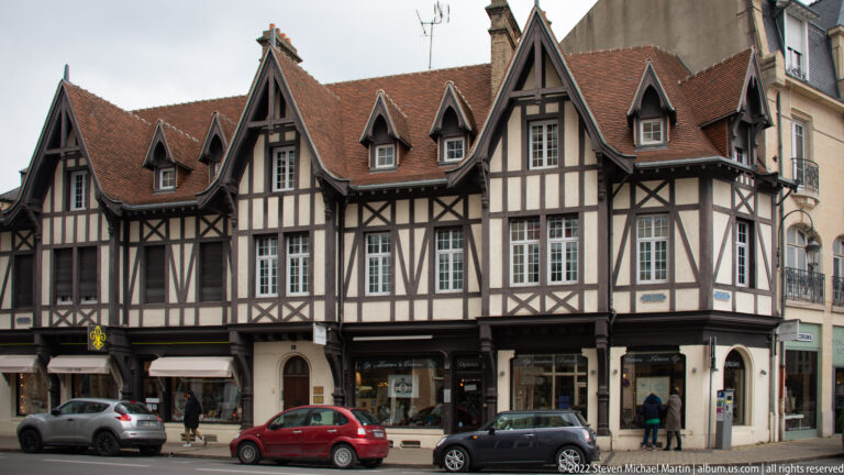 Long half-timbered building in Reims with red tiled roofs, dormers, dark timber framing, and shops at street level.