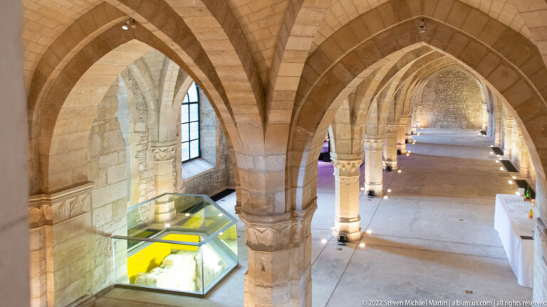 Long stone hall in the Palais du Tau with pointed rib vaults, columns, display cases, and low museum lighting.