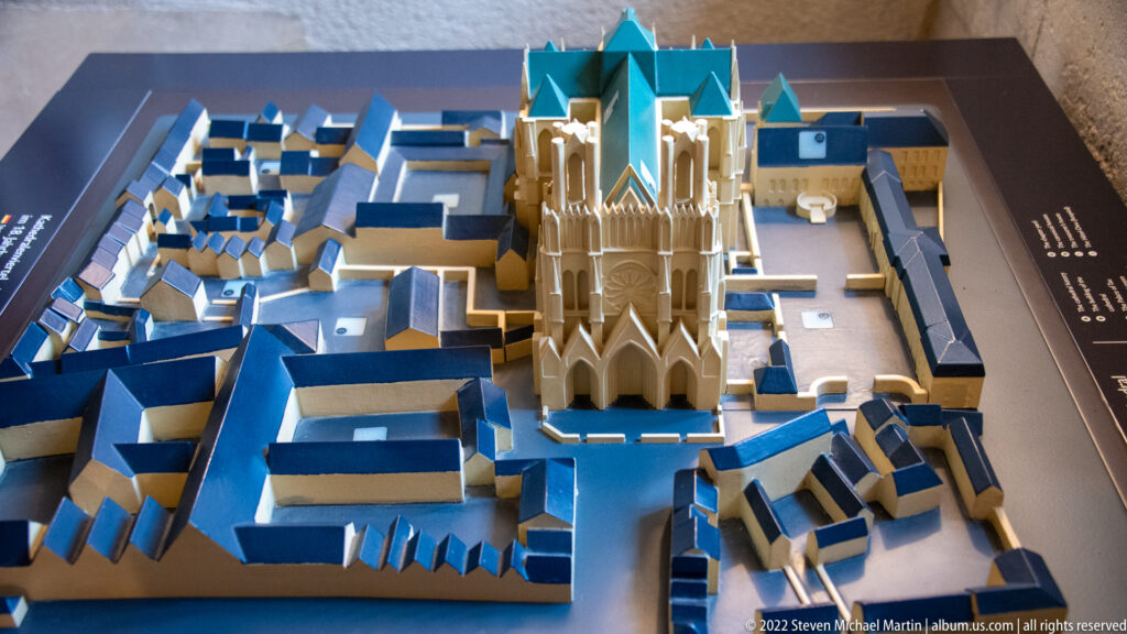 Scale model of Reims Cathedral with twin towers and blue-green roofs surrounded by simplified neighboring buildings.