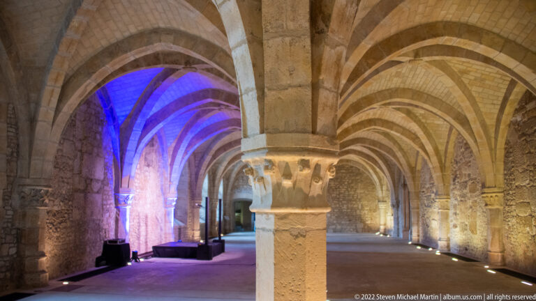 Rib-vaulted stone hall in the Palais du Tau with a central pier, floor lighting, and one bay illuminated blue and magenta.