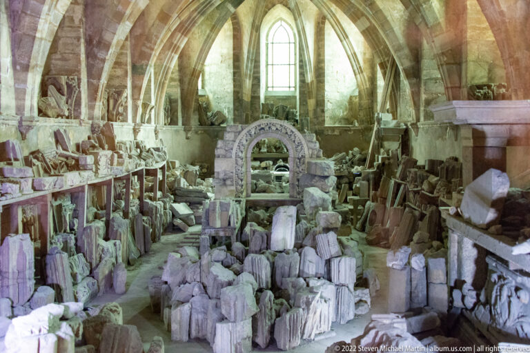 Vaulted museum room densely filled with carved stone architectural fragments, capitals, moldings, and a larger arch element.