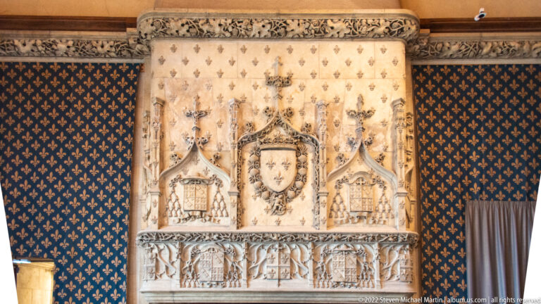 Ornate stone fireplace with a crowned fleur-de-lis shield against blue walls patterned with gold fleurs-de-lis.