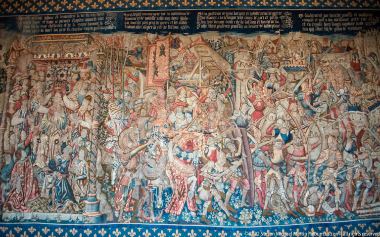 Large historic tapestry showing a crowded battle with horsemen, soldiers, banners, and architecture above a fleur-de-lis border.
