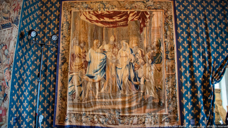 Vertical tapestry showing a ceremonial gathering of richly dressed figures beneath a red canopy, framed by an ornate border.