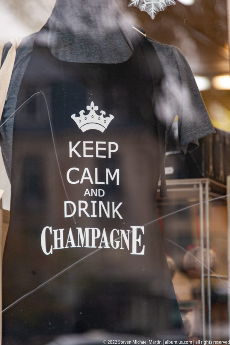 Black T-shirt in a shop window reading “Keep Calm and Drink Champagne,” with reflections visible on the glass.