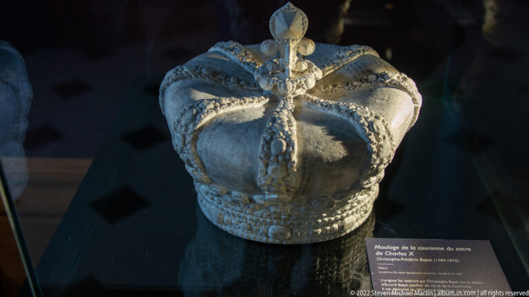 Pale crown mold or model with jeweled bands, fleur-de-lis ornament, and a cross, displayed in a dark museum case.