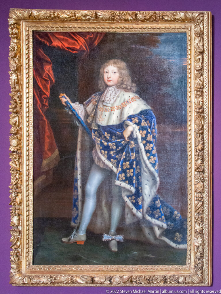 Young royal figure in blue fleur-de-lis mantle and ermine, standing in court dress beside red drapery.