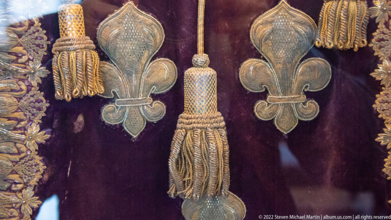 Purple velvet textile with metallic fleur-de-lis embroidery, braided edging, and heavy gold tassels.