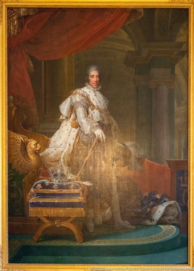 Standing monarch in pale ceremonial robes beside crown-like regalia in an ornate interior.